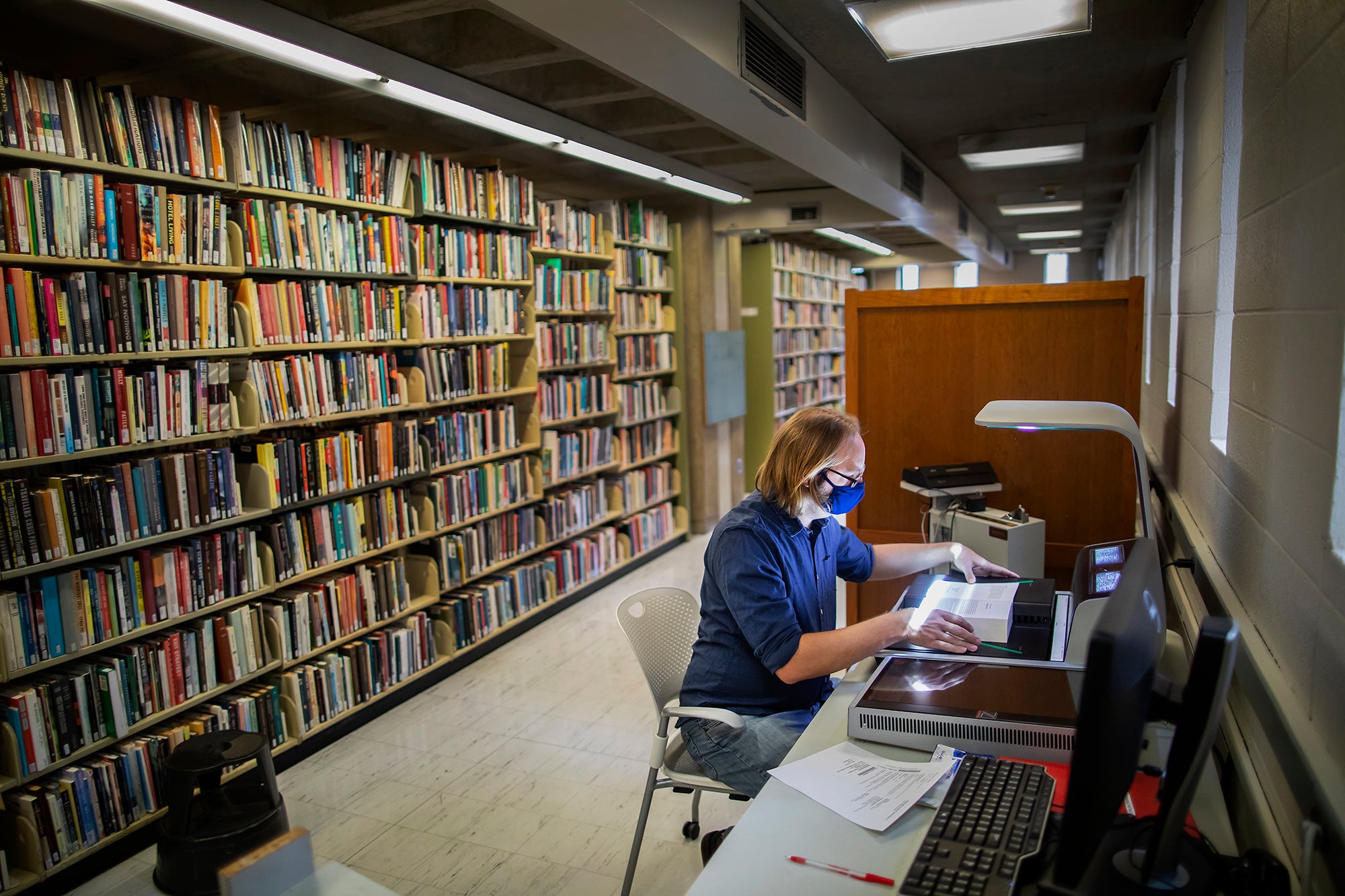 A library staff member sits at a desk scanning pages of a book. Behind him is a floor to ceiling bookshelf.