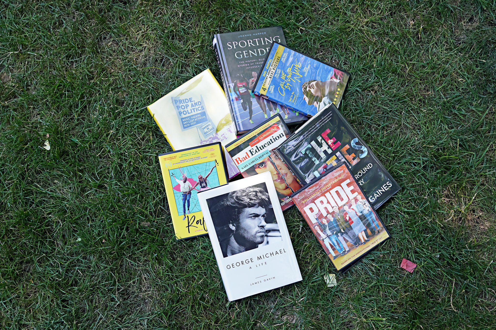 Books and DVDs are laid out in a circular pattern on grass with the DVD Bad Education in the center. Clockwise from the top are the book Sporting Gender, the DVD Call Me By Your Name, DVD Fifties, DVD Pride, book George Michael, DVD Rafiki, and book Pride, Pop and Politics.